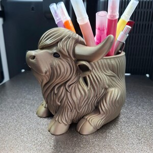 以下が含まれることがあります： さまざまな色のマーカーが入った、金色のハイランド牛の形をしたペン立て。 ペン立ては3Dプリントされており、牛の毛皮、角、ひづめなど、詳細なデザインが施されています。 マーカーの色は、ピンク、オレンジ、黄色など様々です。