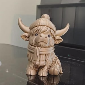 Op de afbeelding: Een goudkleurig beeldje van een Schotse Hooglander, met een wintermuts en sjaal. De koe heeft grote hoorns en een pluizige vacht. Het beeldje zit en heeft een gedetailleerd ontwerp.