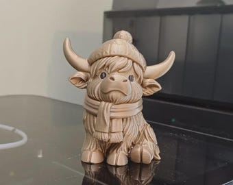 Puede incluir: Figura dorada de una vaca de las Highlands, con gorro y bufanda de invierno. La vaca tiene grandes cuernos y un pelaje esponjoso. La figura está sentada y tiene un diseño detallado.