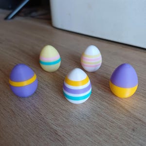 Op de afbeelding: Vijf kleurrijke paasei-decoraties. Elk ei heeft een ander kleurenschema, waaronder paars, geel en wit, met horizontale strepen. De eieren zijn gerangschikt op een houten oppervlak.