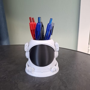 Op de afbeelding: Een witte bureau-organizer in de vorm van een astronautenhelm met pennen. De organizer heeft een groot zwart vizier en gedetailleerde kenmerken. De pennen zijn rood, blauw en zwart.