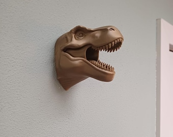 May include: A bronze-colored Tyrannosaurus Rex head sculpture mounted on a light blue wall. The dinosaur head is detailed with sharp teeth and a textured surface, showcasing a prehistoric theme.