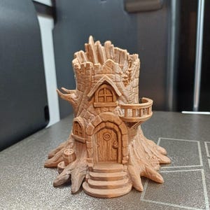 Op de afbeelding: Een gedetailleerd, bruin, 3D-geprint boomhutmodel. De miniatuurstructuur heeft een voordeur, ramen, een balkon en een torenachtig dak. De boomhut staat op een getextureerd oppervlak en toont de details van de schors en architectonische elementen.