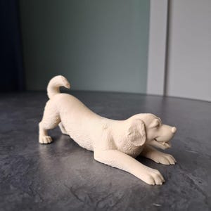 Op de afbeelding: Een crèmekleurig hondenbeeldje in een speelse pose, met uitgestrekte voorpoten en gekrulde staart. De kenmerken van de hond zijn gedetailleerd, tegen een neutrale achtergrond. Dit decoratieve item kan een charmante toevoeging zijn voor elk huis.