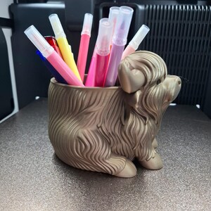 以下が含まれることがあります： ブロンズ色のハイランド牛の形をしたペン立て。カラフルなマーカーで満たされています。マーカーはピンク、赤、黄色、青の様々な色合いです。ペン立ては金属の表面に置かれています。