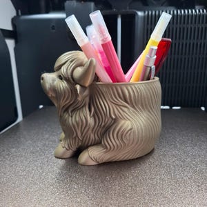 以下が含まれることがあります： さまざまな色のペンやマーカーが入った、金色のハイランド牛型のペン立て。 ペン立ては3Dプリントされており、テクスチャ加工された表面に置かれています。 ペンには、ピンク、赤、黄色、銀色が含まれています。