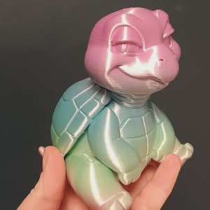 May include: A 3D-printed turtle figurine with a gradient color scheme. The turtle's head is pink, transitioning to blue and green on its shell and body. The figurine has a smiling expression and is held in a hand.