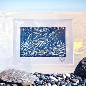 May include: A framed blue and white linocut print depicting an octopus and a bottle in the ocean. The print is in a white frame, displayed on a pebble beach with the sea in the background. The print has the text "Pieces of Eight".