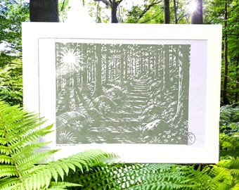 May include: A framed print of a forest scene in shades of green and white. The artwork depicts a path winding through trees, with sunlight shining through the canopy. Large rocks line the path. The print is in a white frame.