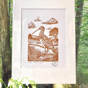 Op de afbeelding: Een ingelijste prent met een bruine linosnede van een wulp in een landelijk landschap, met velden en bomen onder een bewolkte hemel. De prent is gevat in een witte lijst en draagt de tekst "Echoes of the Curlew".