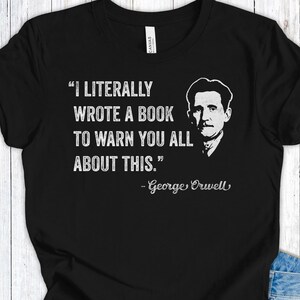 Könnte beinhalten: Schwarzes T-Shirt mit weißem Text und einem Porträt von George Orwell. Der Text lautet: „ICH HABE WIRKLICH EIN BUCH GESCHRIEBEN, UM EUCH ALLE DAVOR ZU WARNEN.“ Der Name des Autors ist ebenfalls enthalten.