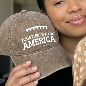 May include: Two baseball caps are displayed. One is brown with the text "TOGETHER WE ARE AMERICA" in white. The other cap has a brown and tan spotted pattern on the underside. The caps are held up.