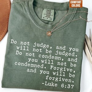 Puede incluir: Camiseta Comfort Colors verde oliva con texto blanco que cita Lucas 6:37. Un collar dorado con un colgante circular descansa sobre la camiseta. La camiseta está doblada y un sombrero marrón es parcialmente visible en el fondo.