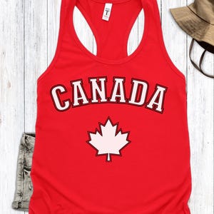 May include: A red tank top with the word "CANADA" in white block letters with a black outline. Below the word is a white maple leaf. The tank top is sleeveless with a racerback design.