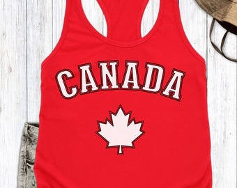 May include: A red tank top with the word "CANADA" in white block letters with a black outline. Below the word is a white maple leaf. The tank top is sleeveless with a racerback design.