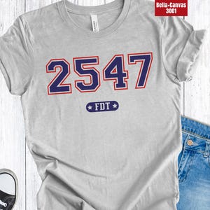Op de afbeelding: Een lichtgrijs t-shirt met het nummer "2547" in marineblauw met een rode omlijning. Onder de cijfers staat de tekst "FDT" in een marineblauwe ster. Het shirt wordt gecombineerd met een blauwe jeans en zwarte sneakers.