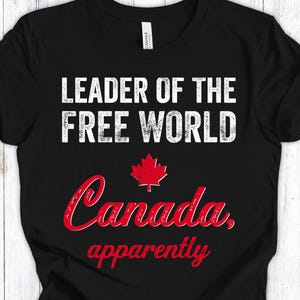 Op de afbeelding: Zwart T-shirt met de witte tekst "LEADER OF THE FREE WORLD". Daaronder een rood esdoornblad en het woord "Canada, apparently" in rode letters.
