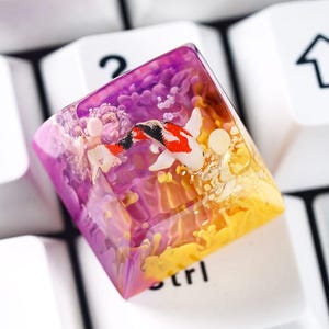 May include: A custom keycap featuring a koi fish design. The keycap is a square shape with a clear resin exterior. The interior design features a koi fish with red, black, and white markings, set against a purple and yellow background.