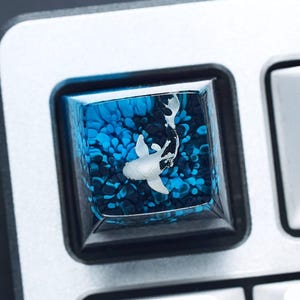 May include: A custom keyboard keycap featuring a koi fish design. The keycap has a clear resin top with a blue and black background, and a white koi fish. The keycap is surrounded by a black frame and is set on a silver keyboard.