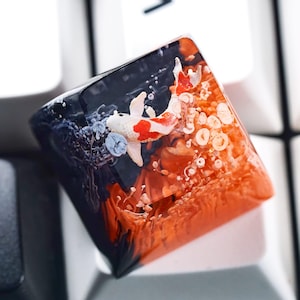May include: A custom keycap featuring a koi fish design. The keycap has a clear resin body with a black and orange gradient, and a white, red, and black koi fish. The keycap is resting on a white keyboard.