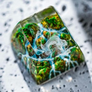 Puede incluir: Una tecla rectangular de resina transparente con un diseño de paisaje en miniatura. El interior presenta terreno verde, amarillo y naranja con detalles en azul y blanco, que se asemeja a una cascada. Un elemento blanco en forma de nube está suspendido dentro de la resina.