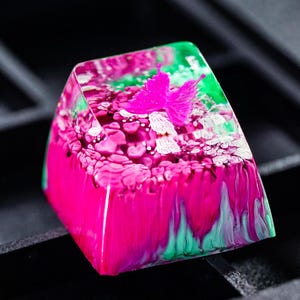 Puede incluir: Una tecla personalizada con un diseño vibrante. La tecla presenta una combinación de colores rosa y verde con un acento de mariposa. La tecla está hecha de resina y está diseñada para teclados mecánicos.