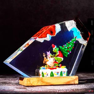 Könnte beinhalten: Ein klarer, kantiger Harzblock mit einer Miniaturszene einer grünen Figur im roten Anzug, die einen geschmückten Weihnachtsbaum hält und neben einem braunen Hund steht. Der Sockel ist ein rechteckiger Holzblock mit einer weiß gemusterten Plattform.