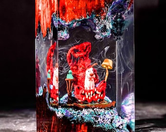 Op de afbeelding: Een rechthoekig sculptuur van hars met een miniatuur witte hond die op een rode stoel zit met een kleine gele lamp. De heldere hars is doordrenkt met wervelende rode, blauwe en zwarte pigmenten, wat een diep, getextureerd en gelaagd effect creëert.