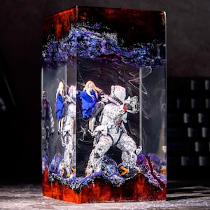 May include: A rectangular resin block containing a miniature astronaut figure in a white suit and a small figure with blonde hair in a blue jacket. The clear resin features swirling white smoke effects, with deep red and purple textured base and top sections.