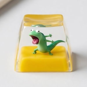 May include: A clear, rectangular keycap with a yellow base and a green lizard figurine inside. The lizard has its mouth open and tongue out. The keycap is on a white surface.