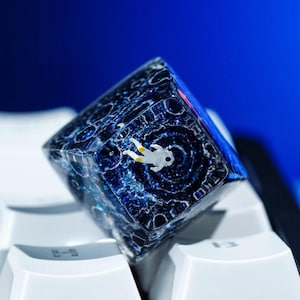 May include: A custom keycap featuring a miniature astronaut floating in a swirling galaxy design. The keycap is a clear cube with a blue and black space-themed interior, set against a white keyboard.
