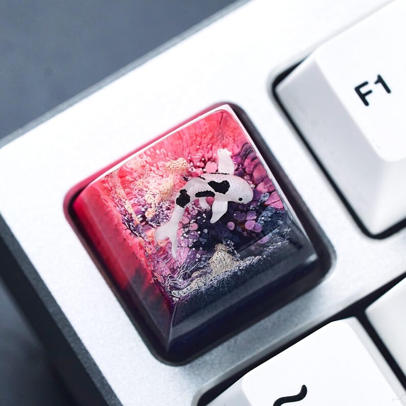May include: A custom keyboard keycap featuring a koi fish design. The keycap has a clear resin body with a vibrant mix of red, pink, purple, and black colors. The koi fish is white with black spots. The keycap is square and sits between other white keys.