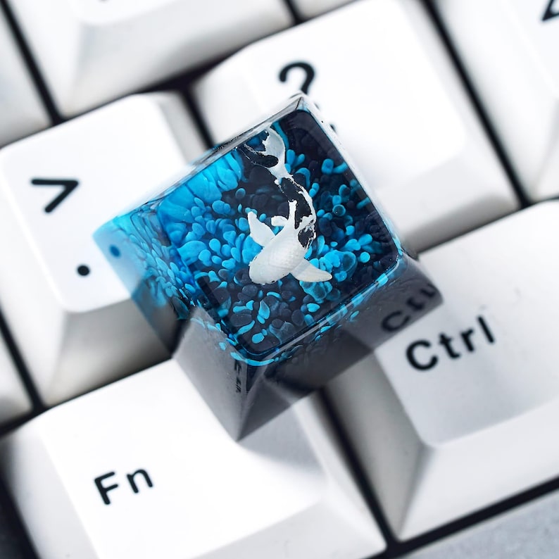 May include: A custom keyboard keycap featuring a koi fish design. The keycap is a square shape with a clear resin top, showcasing a white koi fish swimming in a blue and black background. The keycap is set on a white keyboard.