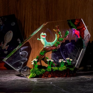 Può includere: Un blocco di resina trasparente contenente una scena in miniatura con una lucertola verde chiaro appollaiata su un ramo d'albero marrone testurizzato. La base presenta muschio verde e piccoli fiori bianchi. Volute di fumo bianco sono sospese.