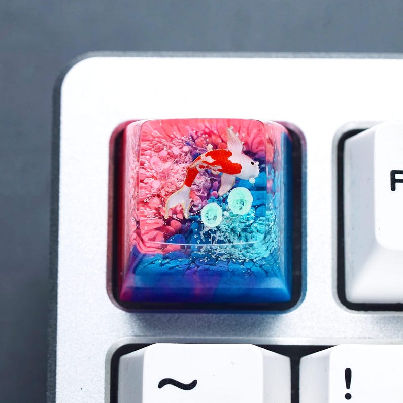 May include: A custom keyboard keycap featuring a koi fish design. The keycap is a square shape with a clear resin exterior. Inside, a red, white, and blue koi fish swims in a colorful, watery environment. The keycap is set on a silver keyboard.