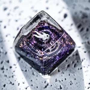 May include: A clear, square-shaped resin piece with a miniature astronaut figure floating within a swirling galaxy of purple and silver glitter. The resin is transparent, allowing light to pass through and highlight the details.