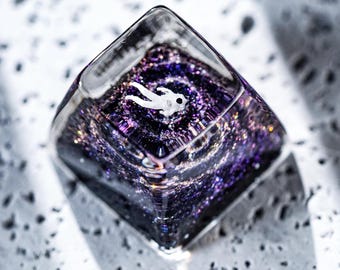 May include: A clear, square-shaped resin piece with a miniature astronaut figure floating within a swirling galaxy of purple and silver glitter. The resin is transparent, allowing light to pass through and highlight the details.