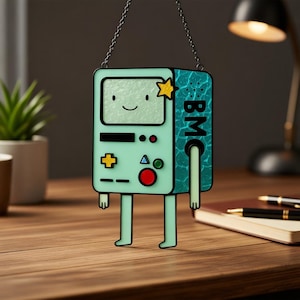 Puede incluir: Un personaje de dibujos animados verde menta con una cara sonriente, una estrella amarilla y las letras "BMC" en un lado. El personaje tiene una pantalla, botones y piernas, y está colgado de una cadena. El fondo está borroso.