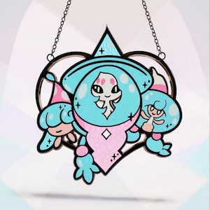 Puede incluir: Un atrapasoles de vidriera con un diseño caprichoso de tres figuras de dibujos animados en tonos azules, rosas y blancos. Las figuras están colocadas sobre un marco negro en forma de corazón, suspendido por una cadena.