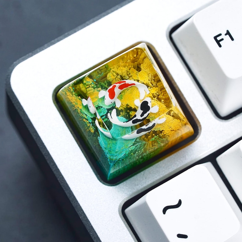 May include: A custom keyboard keycap featuring a koi fish design. The keycap has a square shape with a clear resin top, showcasing a vibrant scene of koi fish in shades of red, black, and white against a yellow and green background. The keycap is set on a silver keyboard.