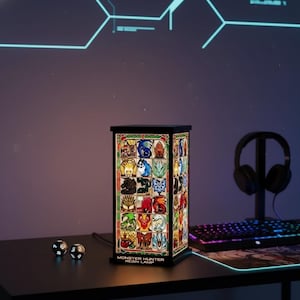 K&ouml;nnte beinhalten: Eine rechteckige Harzlampe mit schwarzem Rahmen, die farbenfrohe Bilder verschiedener Kreaturen zeigt. Die Lampe ist mit "MONSTER HUNTER RESIN LAMP" beschriftet und steht auf einem schwarzen Schreibtisch. W&uuml;rfel und ein Headset sind ebenfalls sichtbar.