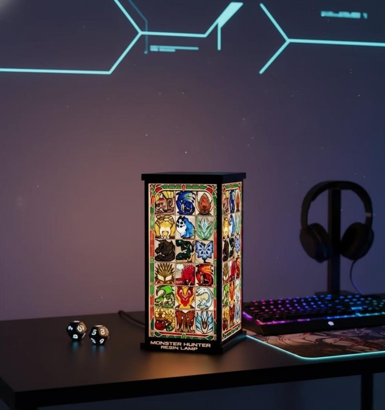K&ouml;nnte beinhalten: Eine rechteckige Harzlampe mit schwarzem Rahmen, die farbenfrohe Bilder verschiedener Kreaturen zeigt. Die Lampe ist mit "MONSTER HUNTER RESIN LAMP" beschriftet und steht auf einem schwarzen Schreibtisch. W&uuml;rfel und ein Headset sind ebenfalls sichtbar.