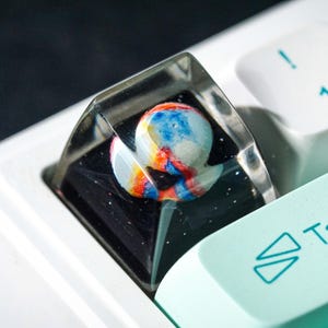 May include: A custom artisan keycap with a colorful, abstract design encased in clear resin. The keycap features a spherical shape with blue, red, and yellow hues, set against a black background. The keycap is on a white keyboard.