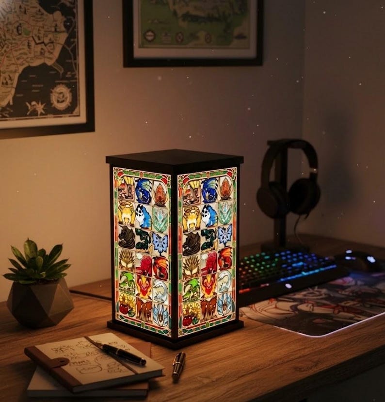 May include: A rectangular stained-glass lamp with a black frame, showcasing colorful fantasy-themed images. The lamp is on a wooden desk, next to a succulent plant, a notebook, and a pen. A keyboard and headphones are also visible.