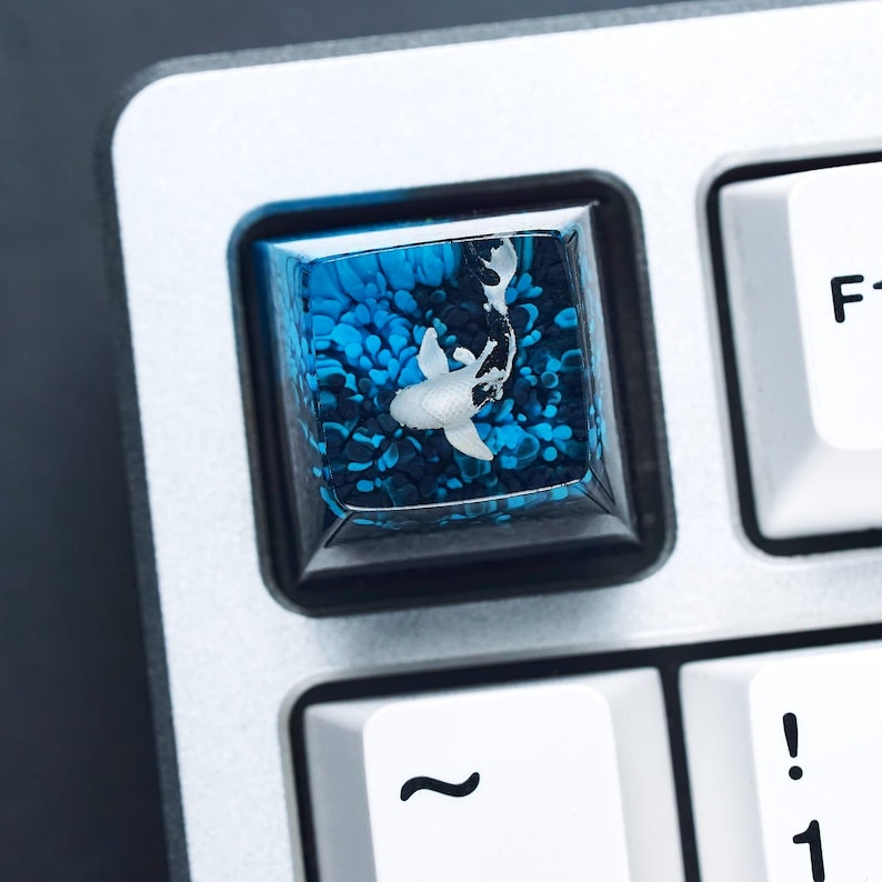 May include: A custom keyboard keycap featuring a koi fish design. The keycap has a clear resin top with a blue and black background, and a white koi fish. The keycap is surrounded by a black frame and is set on a silver keyboard.