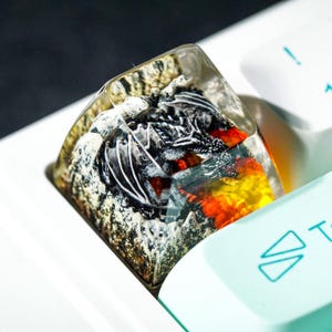 May include: A custom keyboard keycap featuring a detailed dragon design encased in clear resin. The dragon is primarily black and white, with red and yellow accents, set against a rocky, textured base. The keycap is on a white keyboard.