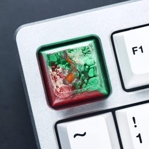 May include: A custom keyboard keycap featuring a miniature orange and white fish swimming in a clear resin enclosure. The keycap has a red and green border, and is set on a silver keyboard.