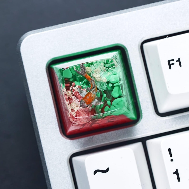 May include: A custom keyboard keycap featuring a miniature orange and white fish swimming in a clear resin enclosure. The keycap has a red and green border, and is set on a silver keyboard.