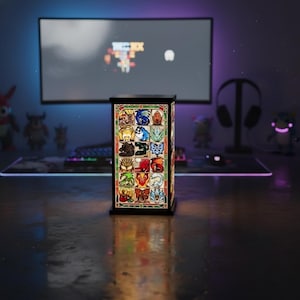 K&ouml;nnte beinhalten: Eine rechteckige Buntglaslampe mit schwarzem Rahmen. Die Lampe zeigt farbenfrohe Bilder verschiedener Kreaturen und Symbole. Die Lampe steht auf einem Schreibtisch mit einem Computermonitor und anderem Gaming-Zubeh&ouml;r im Hintergrund.