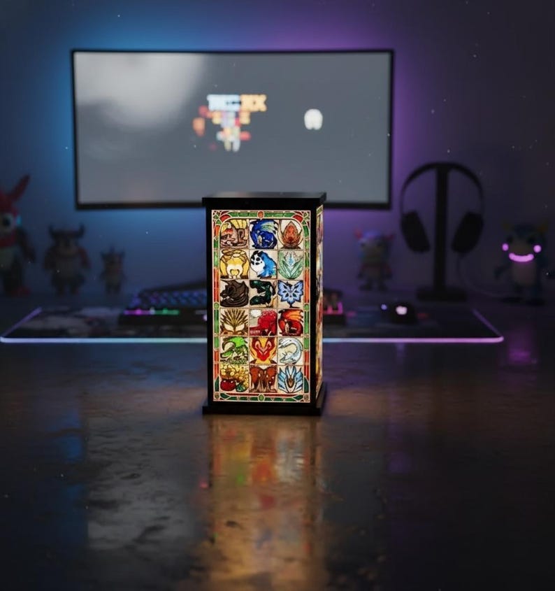 K&ouml;nnte beinhalten: Eine rechteckige Buntglaslampe mit schwarzem Rahmen. Die Lampe zeigt farbenfrohe Bilder verschiedener Kreaturen und Symbole. Die Lampe steht auf einem Schreibtisch mit einem Computermonitor und anderem Gaming-Zubeh&ouml;r im Hintergrund.